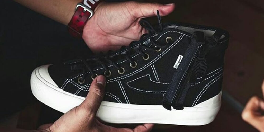 Sepasang tangan memegang sepatu hitam high-cut Compass dengan aksen zipper